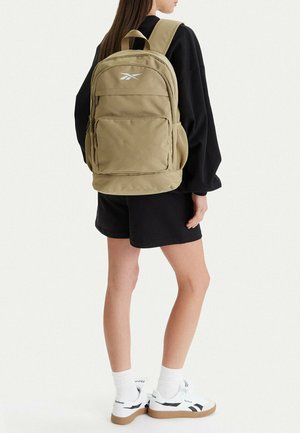 Persona con camisa negra de manga larga y pantalones cortos, llevando una mochila beige de Reebok con bolsillo frontal, combinada con zapatillas blancas de Reebok y calcetines.