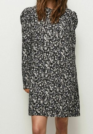 Vrouw in een jurk met zwarte en witte bloemenprint, lange mouwen en pofschouders, staand tegen een egale lichte achtergrond.