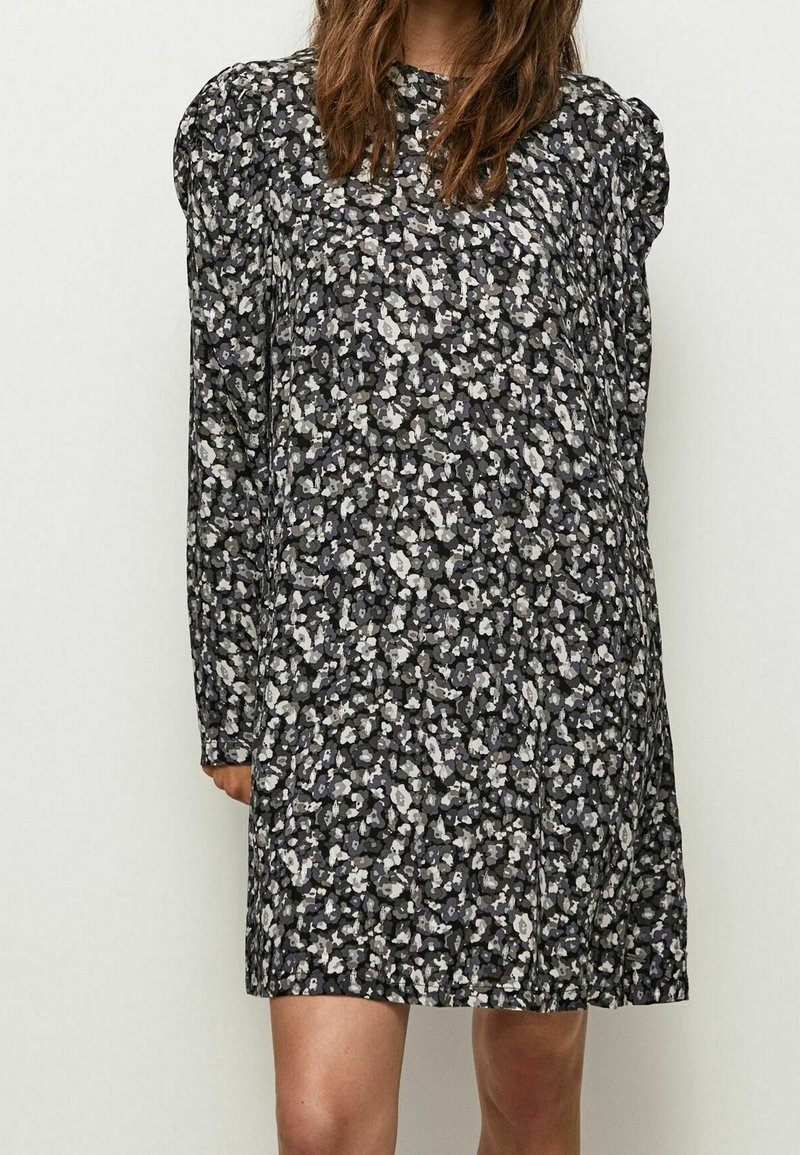Femme portant une robe à manches longues à motif floral noir et blanc avec des épaules bouffantes, debout devant un fond clair uni.