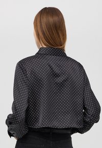 Camicia nera in satin con pois bianchi, design cropped, maniche lunghe e colletto classico. Texture liscia con polsini delle maniche arrotolati.