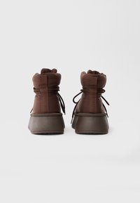 Brune polstrede vinterstøvler med snørebånd, tykke såler og trækkesløjfer, vist fra bagsiden på en ensfarvet hvid baggrund.