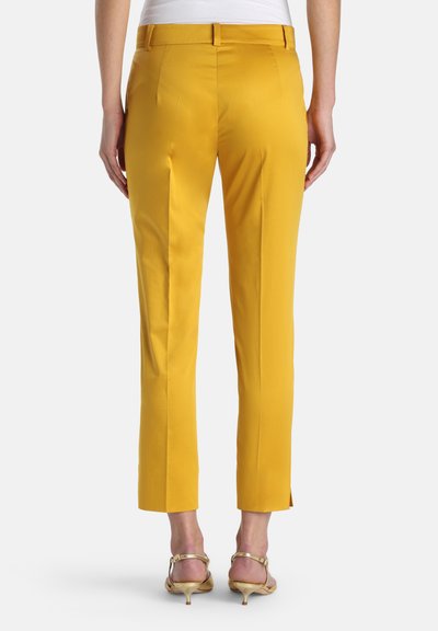 Vue arrière d'une personne portant un pantalon moutarde sur mesure longueur cheville avec des ourlets fendus et des sandales dorées à talons kitten.