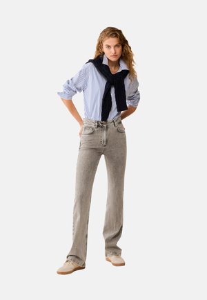 Donna con jeans grigio chiaro a gamba dritta e camicia a righe blu e bianca, con un maglione blu scuro legato intorno alle spalle.