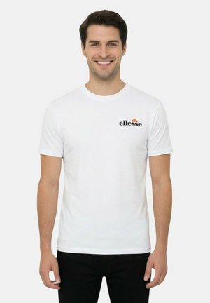 Uomo sorridente che indossa una t-shirt bianca a maniche corte con un piccolo logo "ellesse" sul petto sinistro e pantaloni neri, in piedi.