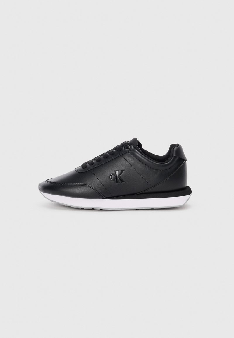 Sneaker in pelle nera con lacci neri, suola bianca e logo "cK" in rilievo sul lato, mostrato di profilo su sfondo bianco.