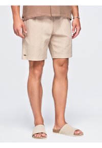 Homme portant un short beige jusqu'aux genoux, un haut marron texturé, un bracelet à perles et des sandales beige ouvertes à l'avant, debout devant un fond uni.