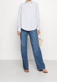 Světle modrá košile na knoflíky s dlouhými rukávy, spárovaná s volnými modrými džíny. Pletená slaměná taška je držena v jedné ruce. Na nohou má ploché sandály.