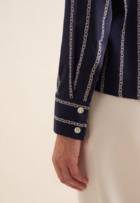 Blusa navy con motivo a catena in bianco e beige, con maniche con bottoni e una texture di tessuto liscia. Primo piano della manica e della mano.