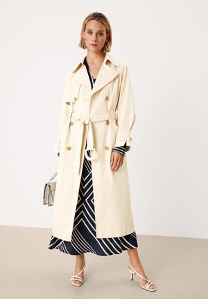 Beige trenchcoat met dubbele rij gouden knopen, strikceintuur, brede revers en grote zakken, gedragen over een marineblauw patroonjurk.