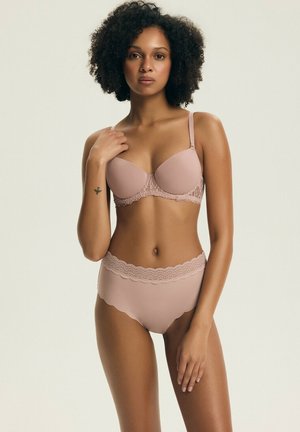 Kobieta o kręconych włosach, ubrana w dopasowany beżowy biustonosz i majtki z wysokim stanem ozdobione koronką, stojąca na tle jasnego, jednolitego tła.