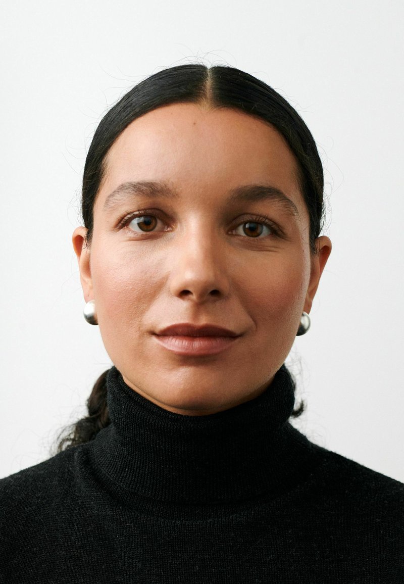 Moteris su rudomis akimis ir tamsiais, atlenktais plaukais, dėvinti sidabro auskarus ir juodą golfą, žvelgianti į priekį priešais paprastą foną.