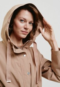 Beżowa wodoodporna kurtka z kapturem, z zamkiem błyskawicznym z przodu, zatrzaskami i tkaninowymi paskami. Gładka tekstura i minimalistyczny design.