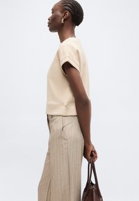 Femme aux cheveux courts tressés, vêtue d’un haut beige à manches courtes et d’un pantalon à fines rayures, tenant un sac à main brun texturé sur le côté, tournée vers la gauche.