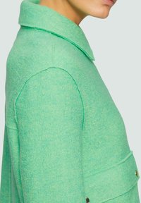 Veste en feutre vert avec un col, des coutures visibles et un détail de poche à l'avant. Le tissu a une apparence texturée et une coupe structurée.