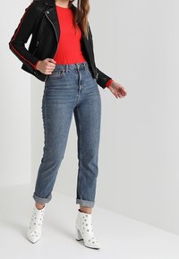 Giacca da motociclista in pelle nera con strisce rosse, maglione a coste rosso, jeans blu a vita alta e stivaletti bianchi con ritagli circolari.