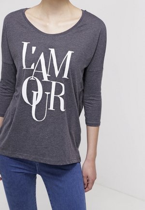 Person trägt ein dunkelgraues Langarmshirt mit weißem stilisiertem Schriftzug "L'AMOUR" und blaue Jeans, steht vor einem einfarbigen hellen Hintergrund.