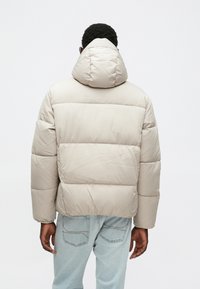 Béžová nadolová bunda s kapucňou, so štepovaným dizajnom a elastickými manžetami. Chrbát je hladký s mierne skráteným strihom.