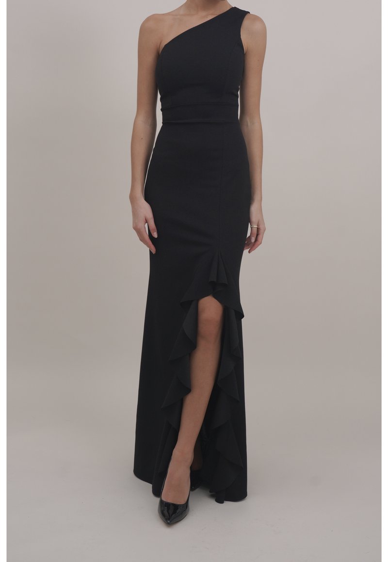 Femme portant une longue robe noire asymétrique avec une fente avant à volants et des talons noirs à bout pointu sur un fond neutre.