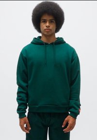 Felpa verde con cappuccio regolabile, polsini e orlo a costine. Realizzata in materiale morbido. Indossata con pantaloni abbinati, caratterizzati da un piccolo logo sul manica.