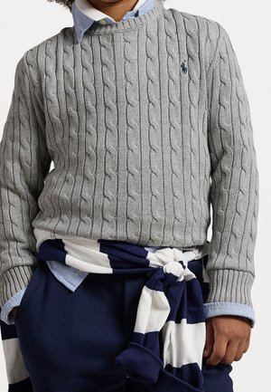 Pull gris en maille torsadée avec poignets et encolure côtelés, porté sur une chemise à col bleu clair ; sweat-shirt rayé bleu marine et blanc noué à la taille.