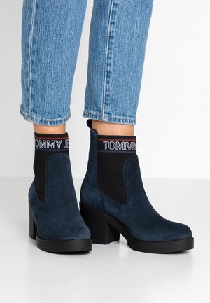 Granatowe zamszowe botki do kostki na słupkowym obcasie, noszone z niebieskimi dżinsami, z czarnymi elastycznymi mankietami z napisem "TOMMY JEANS".