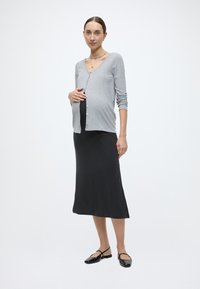 Ζακέτα εγκυμοσύνης σε ανοιχτό γκρι πλεκτό, με ντεκολτέ σε σχήμα V και κουμπιά μπροστά. Φοριέται πάνω από μακρύ, μαύρο, εφαρμοστό φόρεμα και μαύρα επίπεδα παπούτσια.