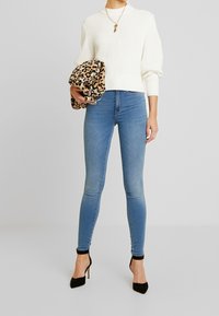 Mujer vistiendo un suéter de punto blanco, jeans ajustados azul claros, tacones altos negros, sosteniendo un bolso de estampado de leopardo contra un fondo liso.