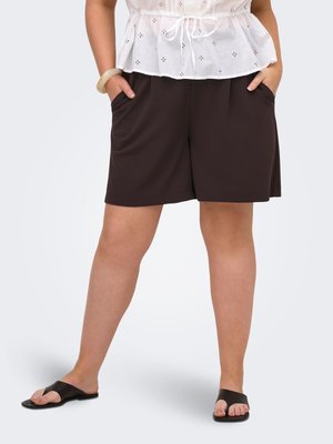 Kvinde iført sorte shorts med vide ben, hvid ærmeløs top med snøre, sorte flade sandaler og et hvidt armbånd, der står med hænderne i lommerne.