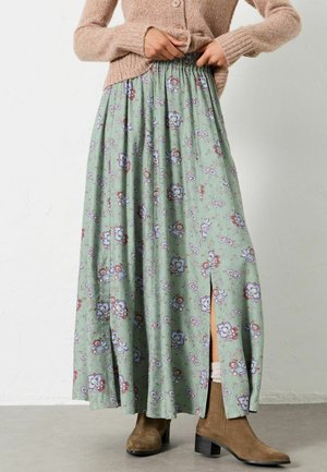 Woman adjusting buttoned beige sweater, wearing green floral maxi skirt with slit and brown suede ankle boots on gray floor.