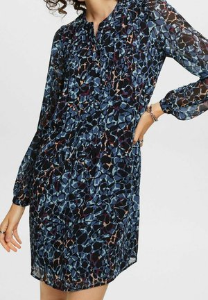 Nainen pukeutunut pitkähihaiseen, polvipituiseen mekkoon, jossa on sininen ja musta abstrakti kuvio, käsi lanteilla, yksiväristä taustaa vasten.