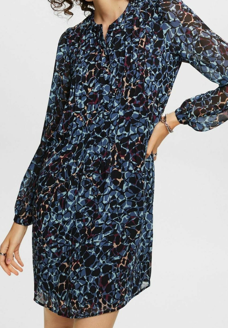 Femme portant une robe à manches longues, longueur genou, avec un motif abstrait bleu et noir, main sur la hanche, sur un fond uni.
