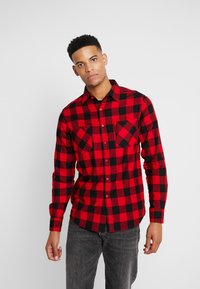 Camisa de franela cuadro rojo y negro con mangas largas, botón al frente y dos bolsillos en el pecho, combinada con jeans grises.