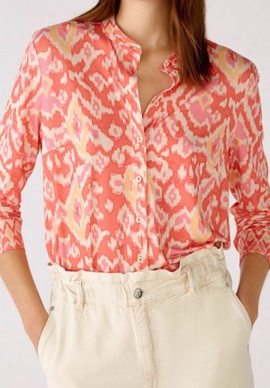 Nainen, jolla on koralli-, vaaleanpunainen ja keltainen kuvioitu napillinen paita neljänneshihoilla, laitettuna korkean vyötärön vaaleanvihreisiin housuihin, kädet taskuissa.