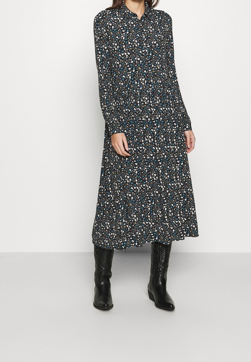 Femme portant une robe midi noire à manches longues avec motif floral, associée à des bottes en cuir noires mi-mollet, debout devant un fond blanc uni.