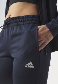 Námornické sportovní kalhoty vyrobené z hladkého materiálu, s elastickým pasem, bočními kapsami a malým stříbrným logem Adidas na levém stehně.