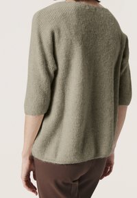 Osoba oblečená v volném, světle šedém pleteném svetru s rukávy sahajícími do loktů a tmavohnědých kalhotách, pohled zezadu.