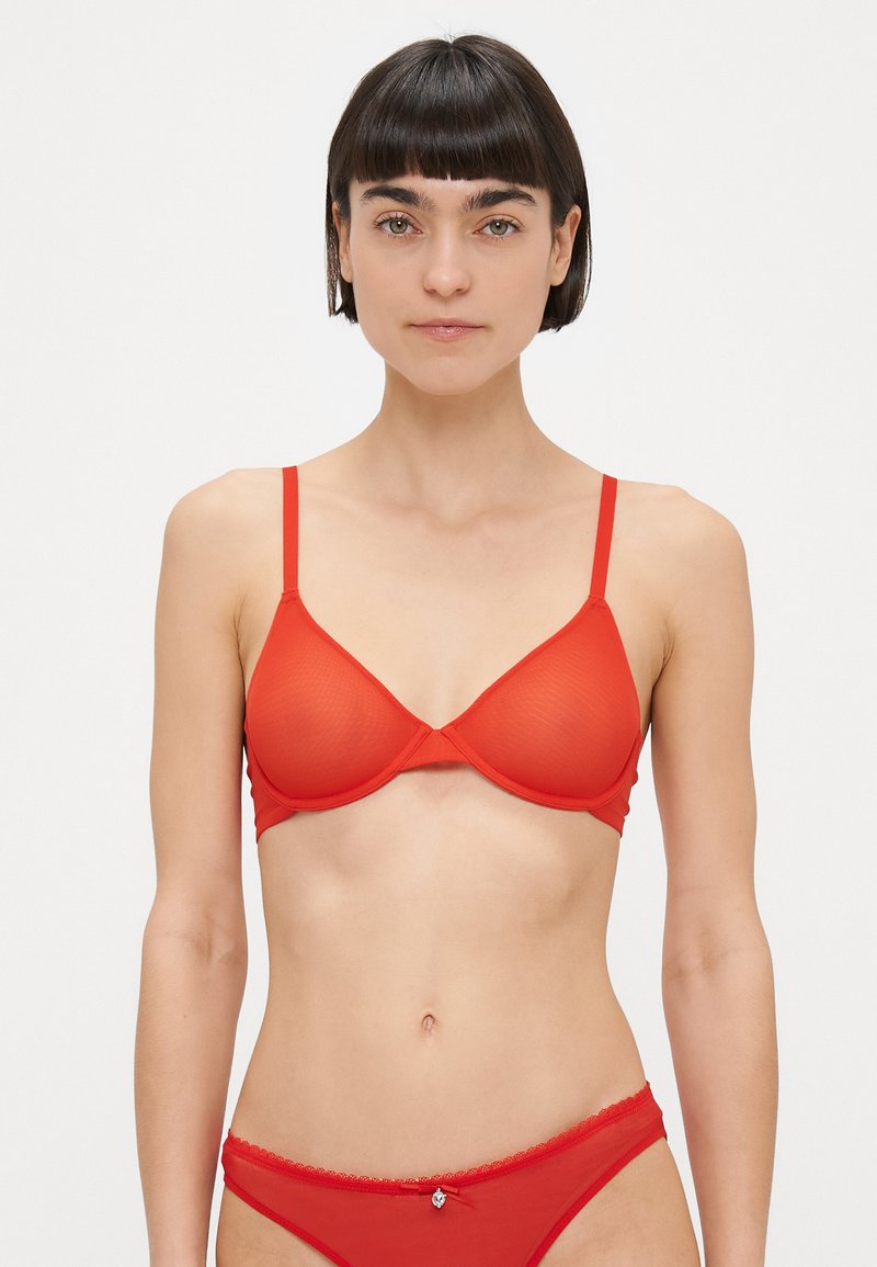 Nő rövid sötét hajjal, piros hálós melltartót és hozzáillő bikini alsóneműt viselve, egy egyszerű fehér háttér előtt áll.