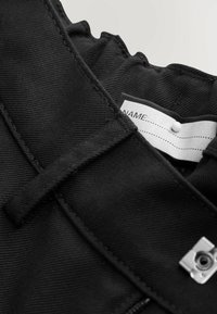 Czarny materiałowy element garderoby o teksturowanym, tkanym wyglądzie, z srebrnym zapięciem na zatrzask i białą metką z miejscem na wpisanie imienia.