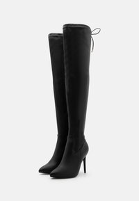 Botas negras de cuero sintético hasta la rodilla con puntera afilada, tacón de aguja y un detalle de cordones único en la parte superior. Textura suave en todo su diseño.