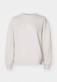 Jasno-beżowa bluza wykonana z mieszanki bawełny, z okrągłym dekoltem, opuszczonymi ramionami oraz białym nadrukiem logo na piersi.