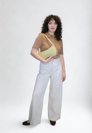 Polo marron à manches courtes, pantalon large blanc, tenant un sac à main jaune incurvé ; fond uni, sans détails supplémentaires.