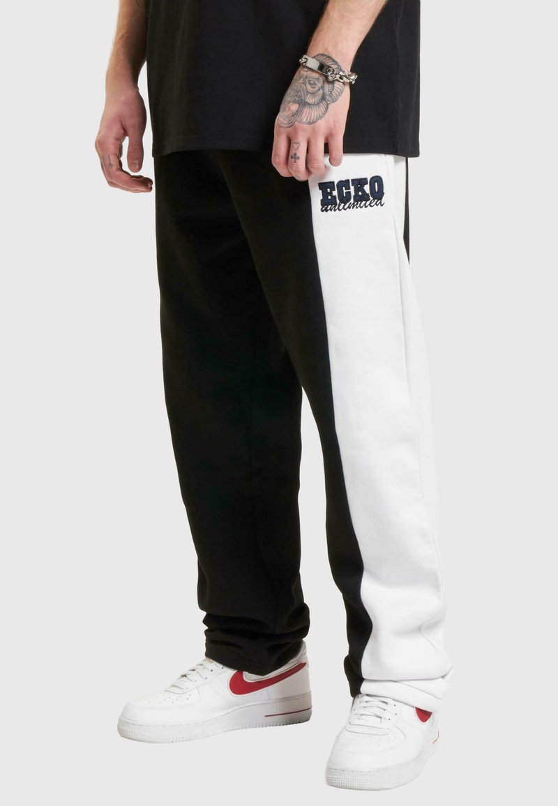 Black and white sweatpants featuring a side stripe, "ECKO" logo on the thigh, cotton material with a relaxed fit, paired with white sneakers.