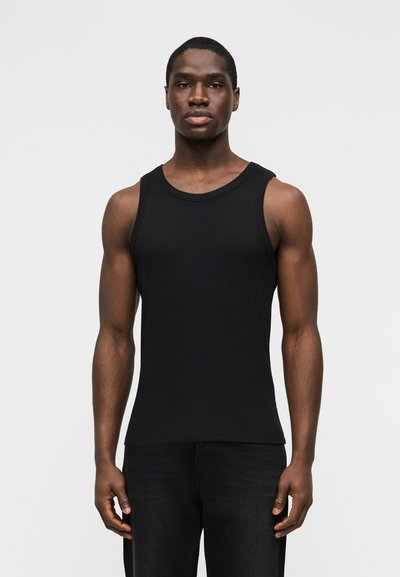 Homme portant un débardeur noir cintré sans manches et un pantalon noir, debout devant un fond blanc uni, face à l'avant.