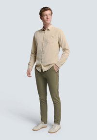 Beige overhemdd met lange mouwen en een borstzak, gecombineerd met olijfgroene slim-fit broek en beige sneakers.