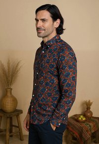 Knoop-up shirt met een donkerblauw bloemenpatroon op een rode achtergrond. De stof is lichtgewicht met lange mouwen en een gestructureerde kraag.