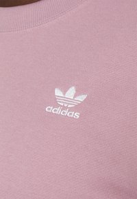 Rosa kortärmad sweatshirt med ribbad rund halsringning och en liten vit broderad Adidas-logotyp på framsidan. Slät bomullstextur.