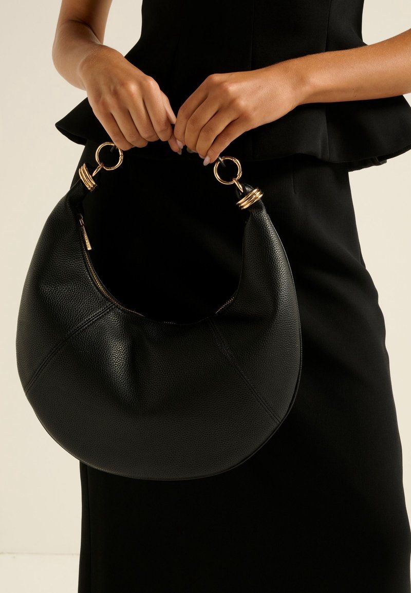 Mains tenant un sac à main en cuir noir en forme de croissant avec des anses en anneaux dorés, porté avec une tenue noire sans manches.