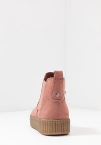 Botín de ante rosa con suela de goma marrón gruesa y tira de agarrar con botón de metal en la parte trasera, mostrado desde atrás sobre fondo blanco.