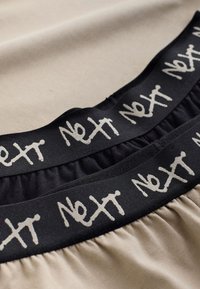 Due paia di fasce elastiche beige e nere con la parola "Next" ripetuta in scrittura bianca su bande nere.