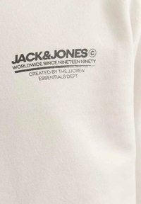 Camiseta blanca con un logo impreso en gris "JACK & JONES" con "MUNDIALMENTE DESDE MIL NOVECIENTOS NOVENTA" debajo y "CREADO POR EL DEPARTAMENTO ESSENTIALS DE JCREW."